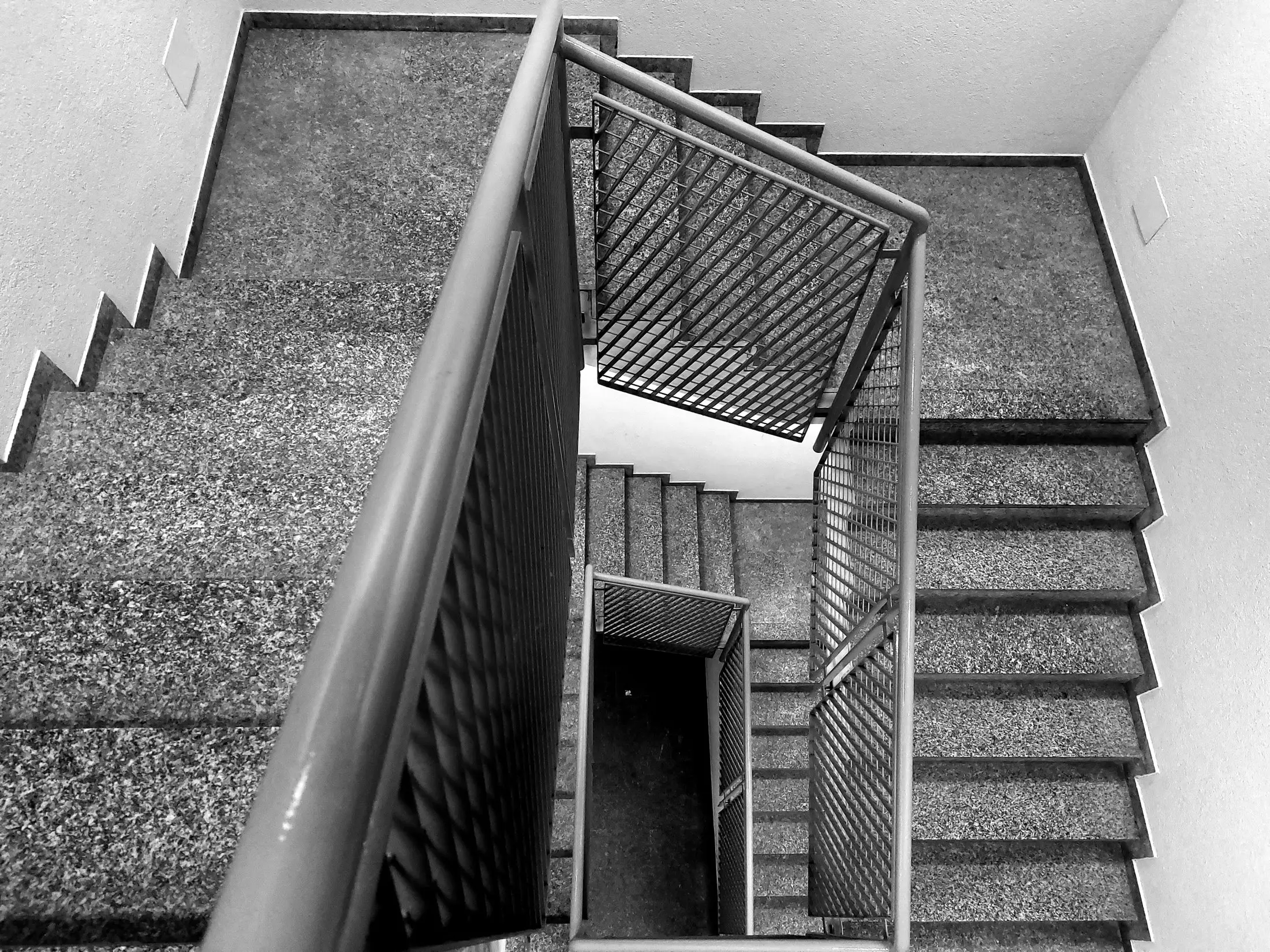 Cage d'escalier vue d'en haut, marches en pierre mouchetée et garde-corps métallique, en noir et blanc