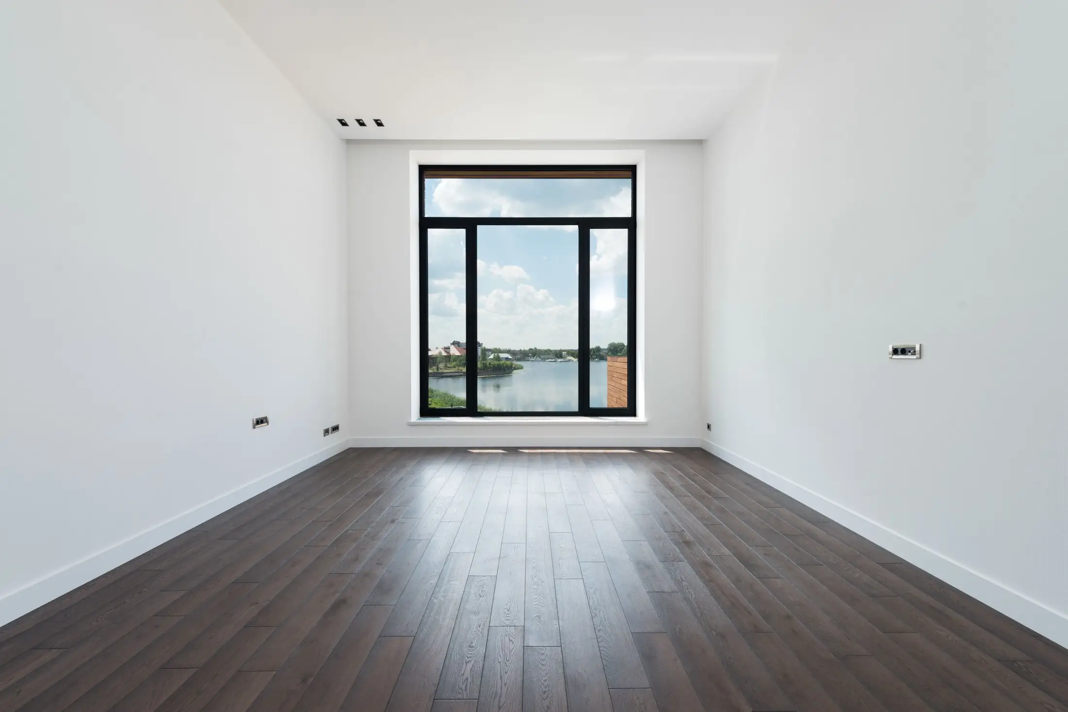 Pièce vide aux murs blancs et au parquet foncé, avec une grande fenêtre donnant sur un plan d'eau