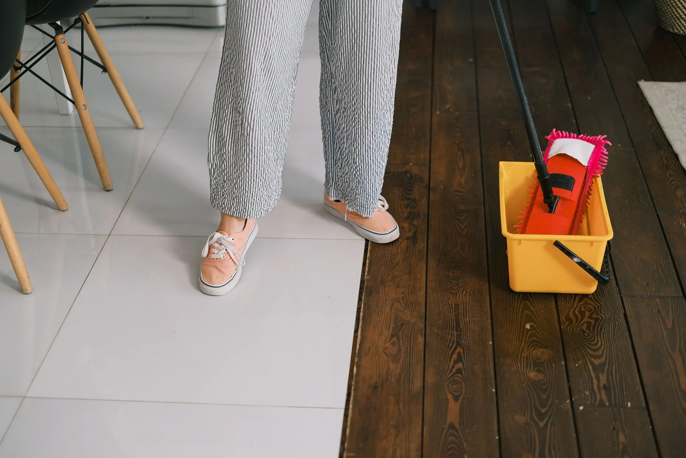 Seau jaune et balai à franges posés entre un carrelage blanc et un plancher foncé